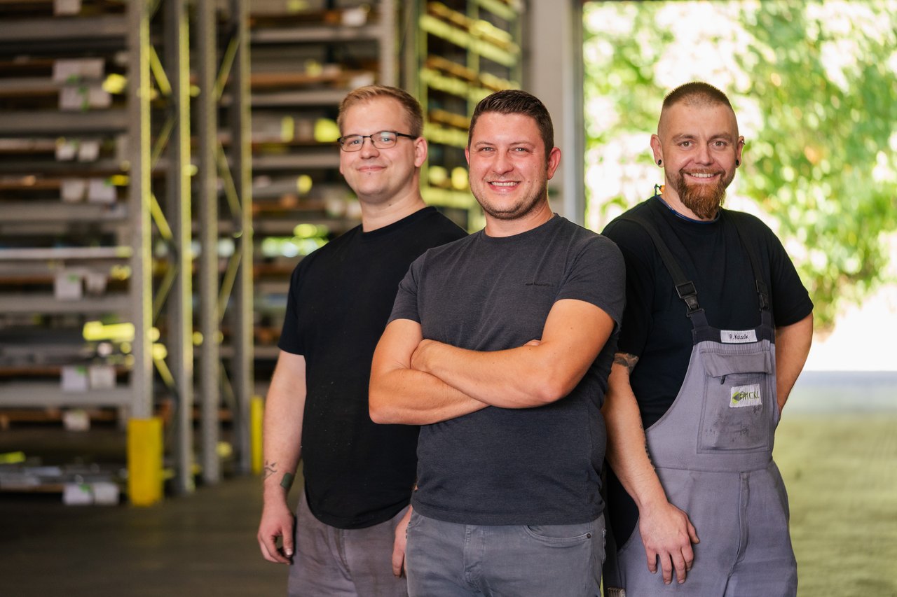 Drei lächelnde Männer in Arbeitskleidung stehen vor Lagerregalen mit Metallprofilen.