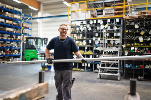 Mitarbeiter trägt lange Metallrohre durch das Lager mit gefüllten Materialregalen.