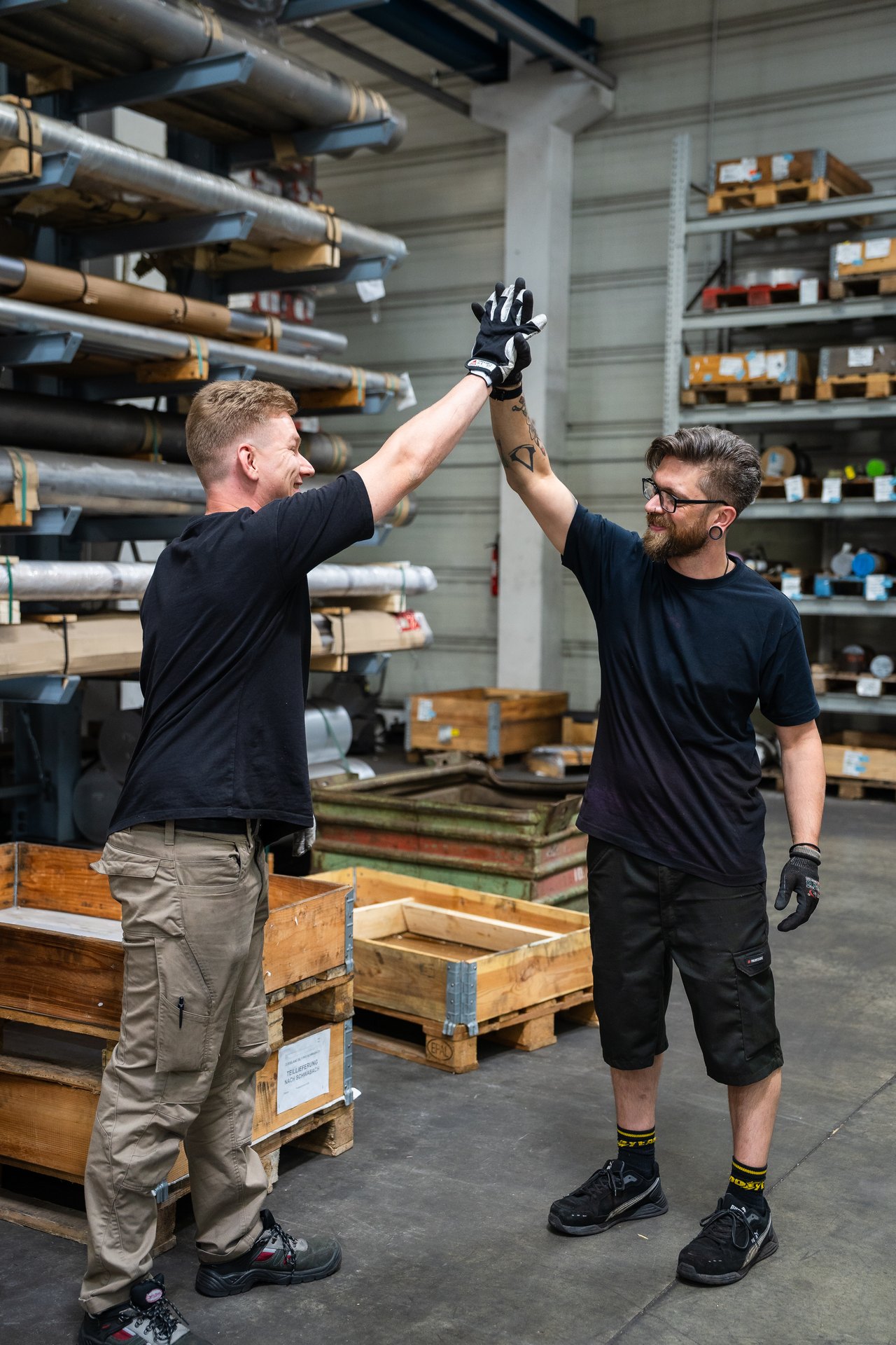 Zwei Männer geben sich in einem Lagerraum mit Metallregalen und Holzkisten ein High-Five.